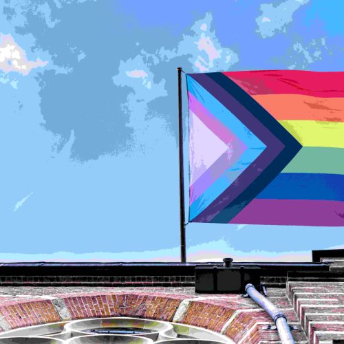 A Progress Pride flag waves atop a brick building against a blue sky with clouds. The flag features rainbow stripes and chevrons in black, brown, light blue, pink, and white.