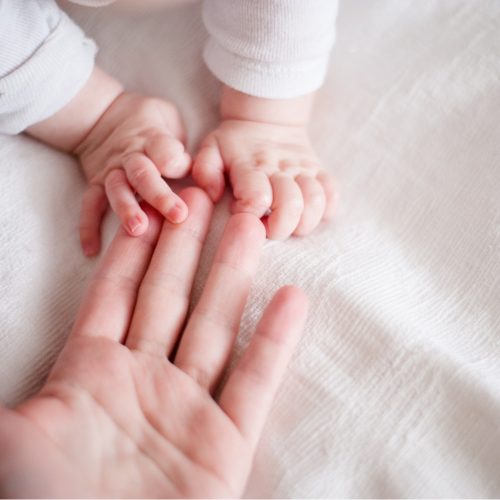 A babys hands gently grasp an adults finger on a soft white blanket. The adults hand is open, and the babys small hands rest on top, creating a tender, intimate moment.