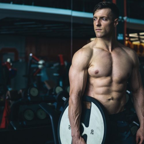 A muscular man stands in a gym holding a large weight plate by his side. He is shirtless, wearing dark workout shorts, and looking to the side. Gym equipment and mirrors are visible in the background.
