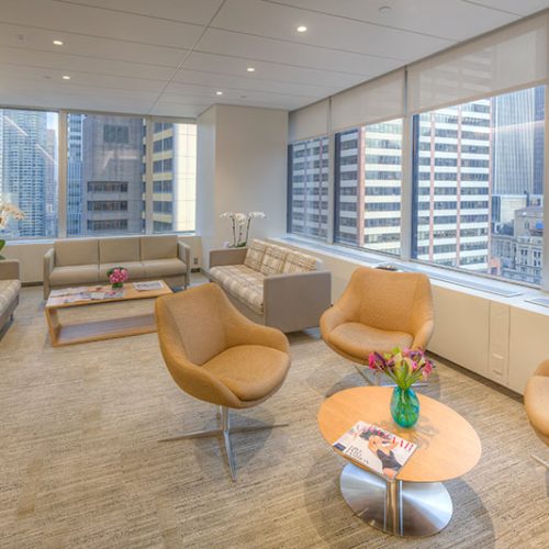 Modern office lounge with large windows showing city skyscrapers. The room has beige armchairs, two sofas, a coffee table with magazines and flowers, and neutral-toned carpet, creating a bright, welcoming atmosphere.