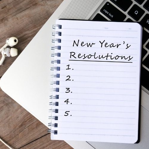 A notebook with New Years Resolutions and a numbered list from 1 to 5 sits on a laptop next to earphones and a green pen on a wooden surface.