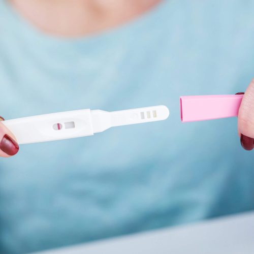 A person holding a pregnancy test with a single line in the result window, indicating a negative result. The person is wearing a light blue top and has red nail polish.