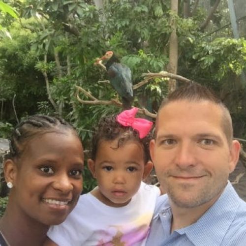 A smiling woman, a man, and a young girl with a pink bow pose for a selfie outdoors, surrounded by lush green plants, with a bird perched on a branch behind them.
