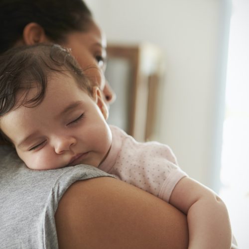 A baby sleeps peacefully on an adult’s shoulder, with the adult facing away and gently holding the child in a cozy, softly lit room.