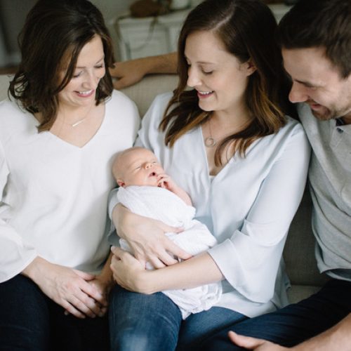 Three adults sit close together on a couch, smiling and looking at a newborn baby wrapped in a white blanket, held by the woman in the center. The atmosphere is warm and joyful.