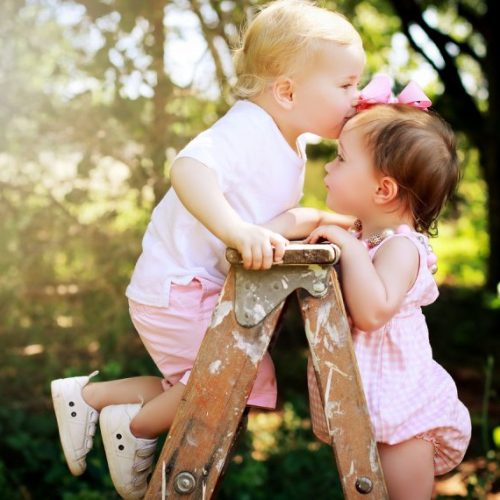 Two young children in pink clothes stand on a wooden ladder outdoors. One child kisses the other on the forehead, both smiling, with green trees and sunlight in the background.