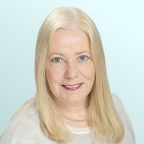 Smiling woman with long straight blonde hair wearing a light gray sweater and a pearl necklace, posed against a light blue background.