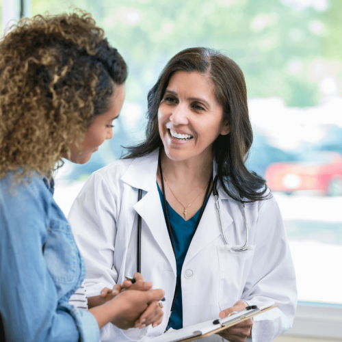 A smiling doctor in a white coat talks with a patient, holding a clipboard. They are standing indoors near large windows with sunlight coming in. The patient listens attentively and both appear engaged in conversation.