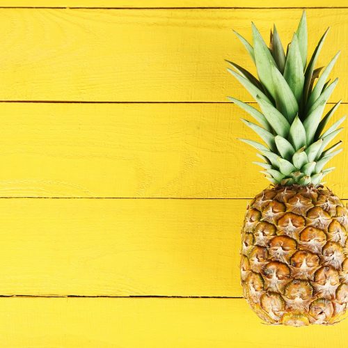 A whole pineapple with green leaves sits on the right side of a bright yellow wooden background.