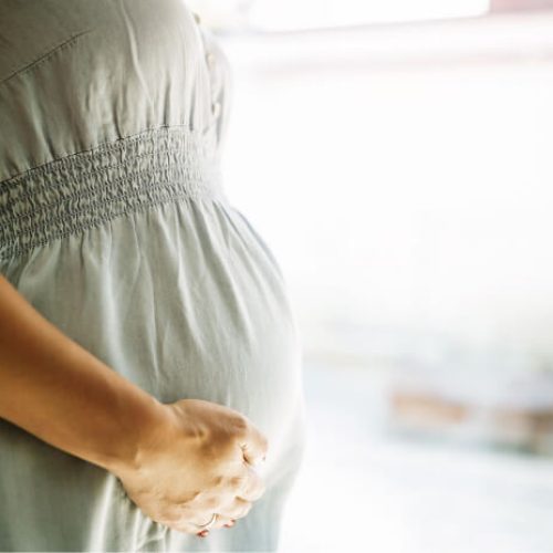 A close-up of a pregnant woman wearing a light gray dress, gently holding her belly with one hand, standing indoors with a soft background.