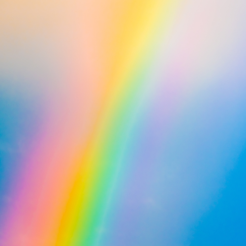 A vibrant rainbow with distinct colors arcs across a soft blue sky, blending into light, wispy clouds. The colors are bright and vivid, creating a cheerful and uplifting atmosphere.
