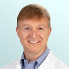 A smiling man with light brown hair wearing a white lab coat and a light blue collared shirt, posed in front of a light blue gradient background.