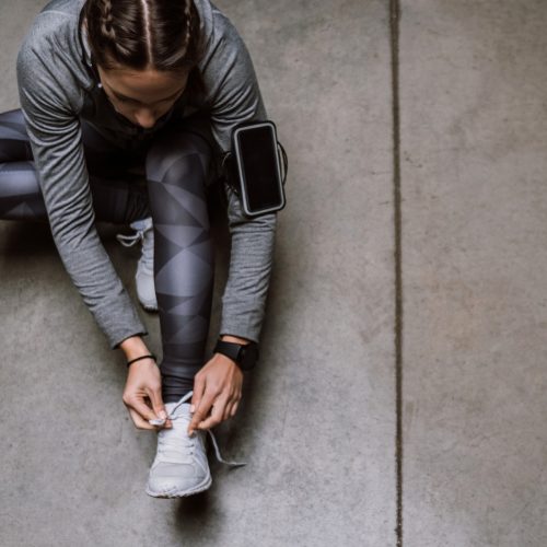 A person in athletic wear sits on the ground tying their shoe. They have a smartphone strapped to their upper arm and wear a smartwatch. The background is a smooth concrete floor.