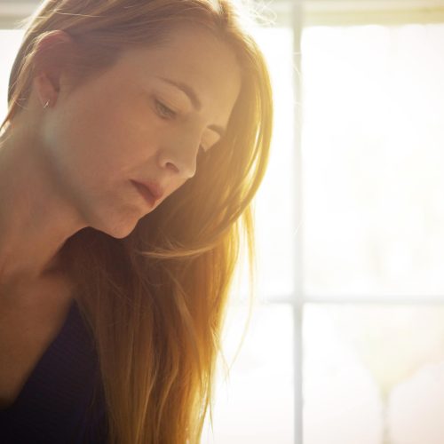 A woman with long red hair looks down thoughtfully, softly lit by sunlight coming through a window behind her. She wears a dark top, and the background is blurred and bright.