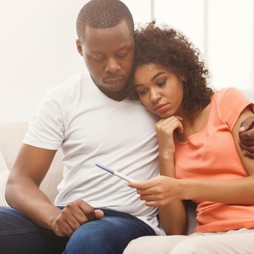 A couple sits closely on a couch, looking concerned as they hold a pregnancy test together. The woman leans on the man’s shoulder, and he has his arm around her for comfort.