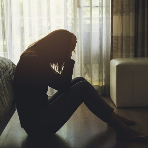 A person sits on the floor by a bed, silhouetted in natural light from a window, holding their head in their hands, appearing distressed or upset in a dimly lit room.
