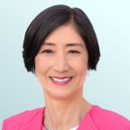 A woman with short black hair wearing a bright pink blazer and a silver necklace smiles at the camera against a light blue background.