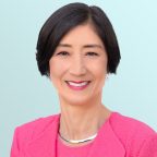 A woman with short black hair wearing a bright pink blazer and a silver necklace smiles at the camera against a light blue background.