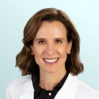 A woman with short brown hair smiles at the camera, wearing a white lab coat over a black shirt. She has stud earrings, and the background is a light blue gradient.