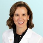 A woman with short brown hair smiles at the camera, wearing a white lab coat over a black shirt. She has stud earrings, and the background is a light blue gradient.