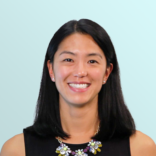 A woman with straight black hair and a floral necklace, wearing a sleeveless black top, smiles in front of a light blue background.