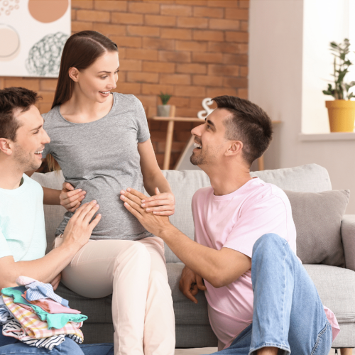 Two men sit by a pregnant woman on a couch, smiling and touching her belly. The happy group relaxes in a cozy living room with baby clothes and plants, where they chat about modern decor and the benefits of Vitamin D for fertility.