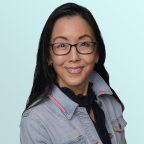 A woman with long dark hair and glasses smiles at the camera. She is wearing a light denim jacket over a black top, standing against a light blue background.