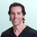A man with dark wavy hair and light stubble smiles at the camera. He is wearing a black V-neck shirt and is posed in front of a light blue background.