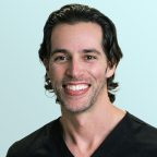 A man with dark wavy hair and light stubble smiles at the camera. He is wearing a black V-neck shirt and is posed in front of a light blue background.