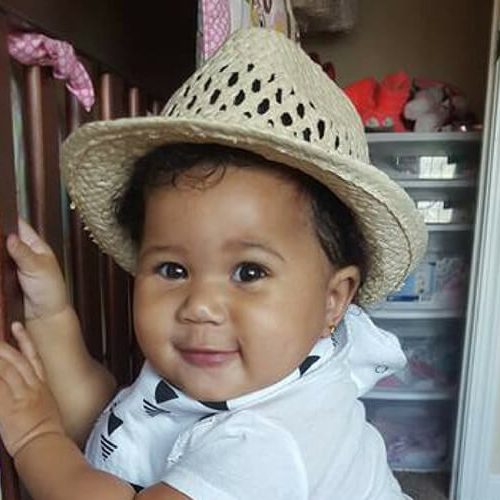 A smiling baby wearing a straw hat and a white outfit stands holding onto a crib rail in a cozy, well-lit room.