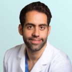 A man with short dark hair and a beard, wearing a white medical coat, smiles at the camera against a light blue background.