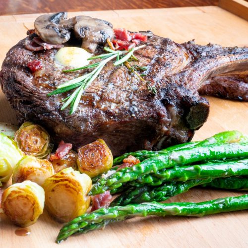 A grilled ribeye steak topped with mushrooms and rosemary sits on a wooden board, served with roasted Brussels sprouts, bacon pieces, and sautéed asparagus.