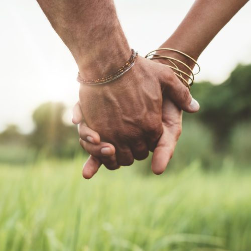 Close-up of two people holding hands outdoors, with one person wearing bracelets. Green grass and trees are visible in the blurred background, and sunlight shines softly between them.