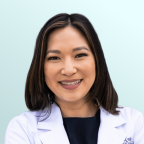 A woman with medium-length dark hair smiles at the camera. She is wearing a maroon top and a delicate necklace with a pendant. The background is a light blue gradient.