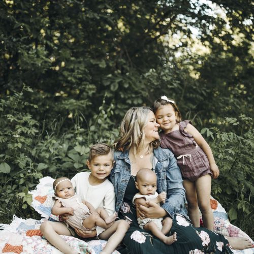 A woman sits on a quilt outdoors with four young children—two babies in her lap, a toddler girl standing and hugging her, and a young boy sitting close—surrounded by lush greenery.