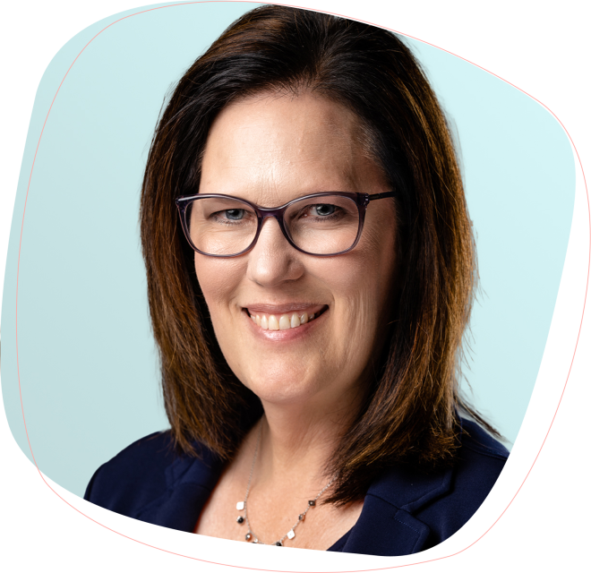 A woman with shoulder-length brown hair and glasses smiles at the camera. She is wearing a navy blazer and a delicate necklace, with a light blue background behind her.