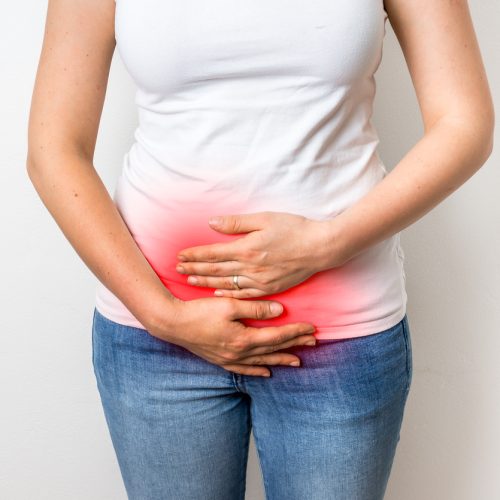 A person wearing a white shirt and blue jeans holds their lower abdomen with both hands; a red highlight on the area suggests pain or discomfort.