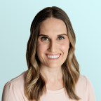 A woman with long, wavy brown hair and light highlights smiles at the camera. She is wearing a light pink top and is posed in front of a pale blue background.