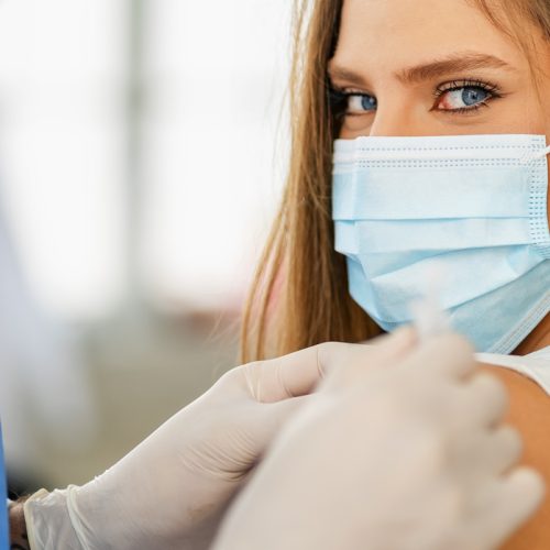 A person wearing a surgical mask receives a vaccine injection in their upper arm from a healthcare worker in protective gloves and a blue gown. The person looks directly at the camera.