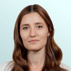 A woman with long brown hair wearing a white coat and a green blouse stands against a light blue background, looking calmly at the camera.