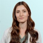 A woman with long brown hair wearing a white coat and a green blouse stands against a light blue background, looking calmly at the camera.