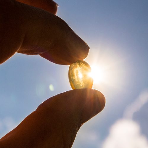 A hand holds a translucent capsule up to the sky, with sunlight shining through it and clouds in the background.