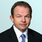A middle-aged man with short brown hair, wearing a black suit, white shirt, and blue patterned tie, poses against a light blue gradient background.