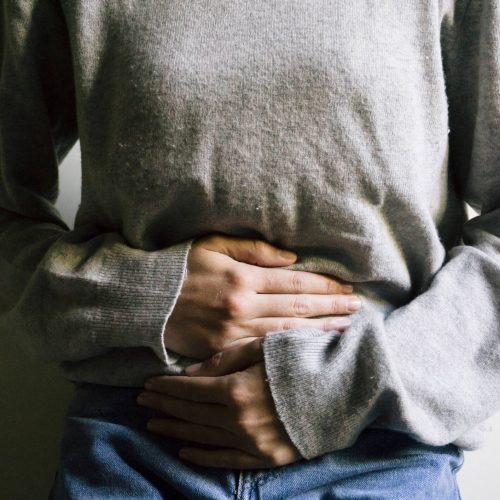 A person wearing a gray long-sleeve shirt and blue jeans holds their hands over their stomach, suggesting discomfort or pain.