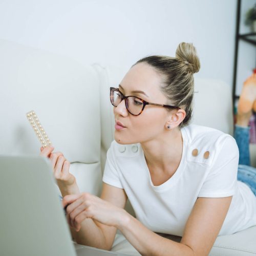 A woman wearing glasses lies on a couch, looking at a pack of pills in one hand while using a laptop with the other hand.