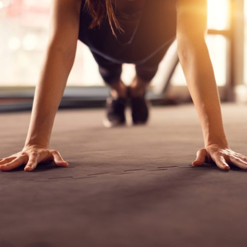 A person in athletic wear performing a push-up on a gym floor with their hands spread wide, focused on exercise. Sunlight streams in from windows in the background.