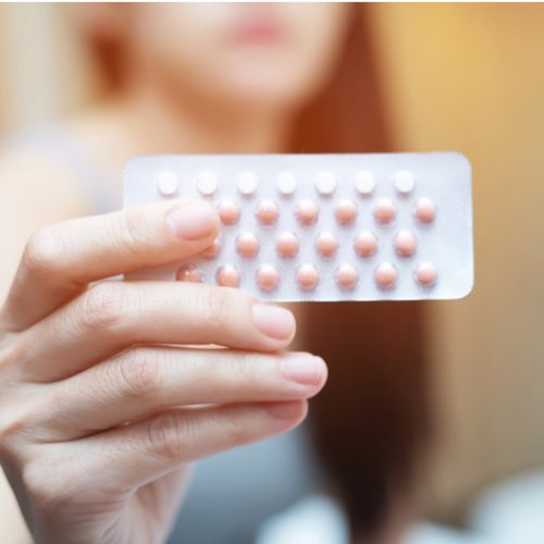 A person holds a blister pack of oral contraceptive pills, with pink and white tablets visible, against a blurred indoor background.