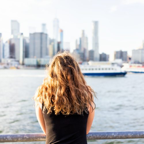 A person with curly hair, seen from behind, stands by a railing overlooking a river with a city skyline and a boat in the background on a sunny day.