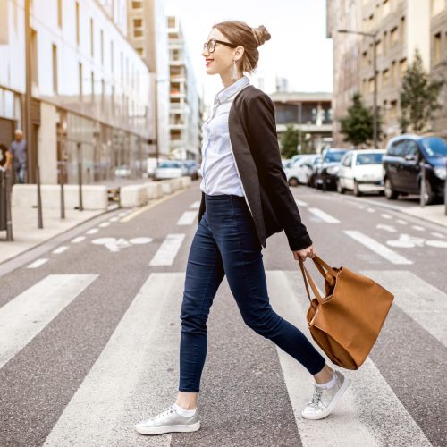 A woman with glasses walks confidently across a city crosswalk, carrying a large brown handbag and wearing a black blazer, white shirt, blue jeans, and white sneakers. Buildings and parked cars line the street.
