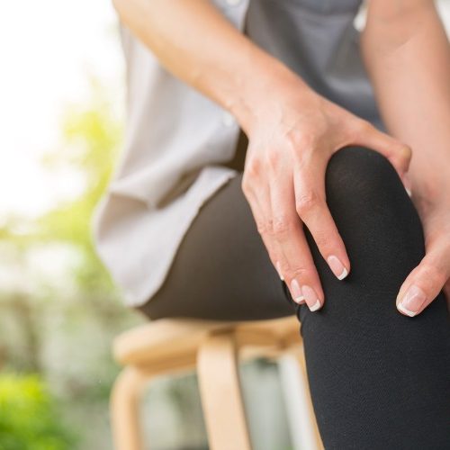 A person sitting on a wooden stool holds their knee with both hands, appearing to experience knee pain or discomfort. The background is softly blurred with natural light.